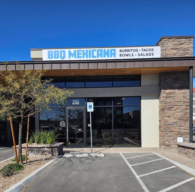 Daytime photo of the exterior of the new BBQ Mexicana restaurant on Sunset Road near Durango in Southwest Las Vegas. The sign above their strip mall location reads "BBQ Mexicana" in light blue, Mexican-style script. To the right of their restaurant name in black letters the sign reads "Burritos, Tacos, Bowls, Salads." There are tinted glass windows on the facade of the restaurant. A tinted glass entrance door shows the suite number 200 above the door. The parking space in the photo shows a handicapped parking sign at the head of the space. To the right of the location is a stone facade and low wall. To the left of the location is a median with a tree and various green leafy plants. It is a sunny day with no clouds in the photo.