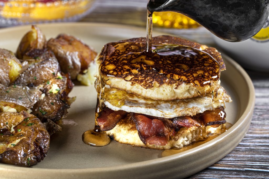 A fried egg and bacon is sandwiched between two pancakes that are being covered with syrup. They are served next to hash browns on a gray plate.