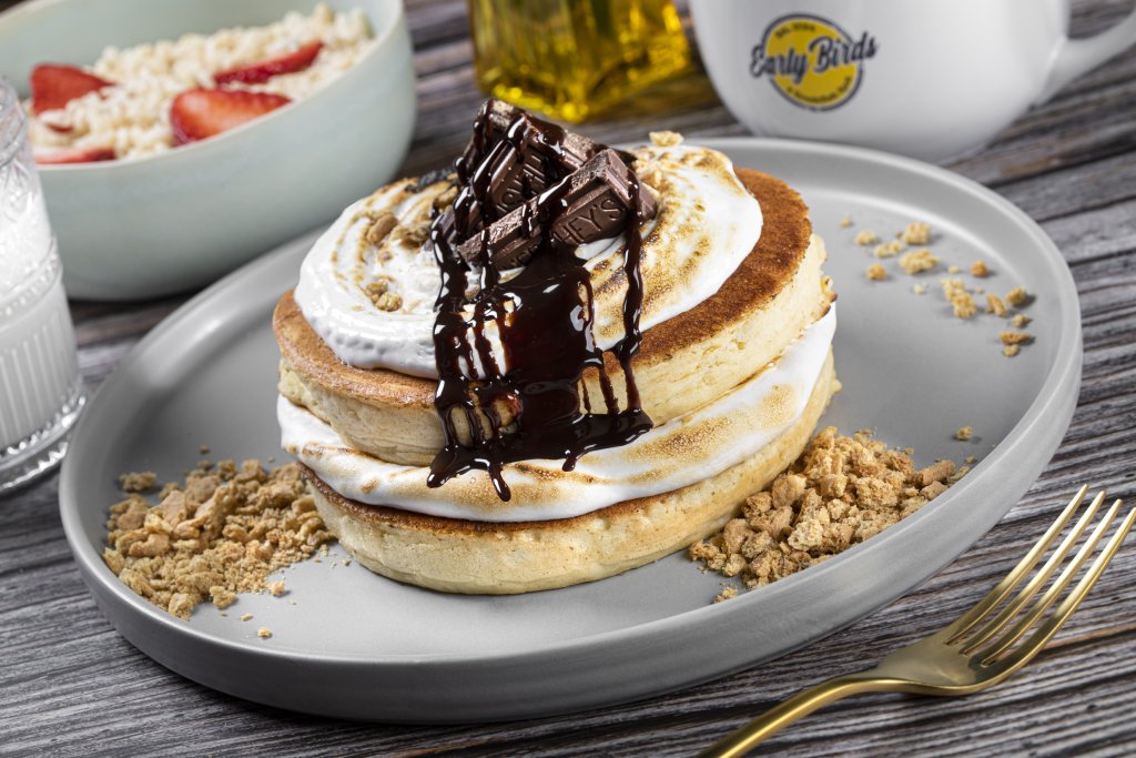 A stack of s'mores pancakes are served on a gray plastic plate. They are covered in whipped cream, chocolate syrup and a hershey mini bar.