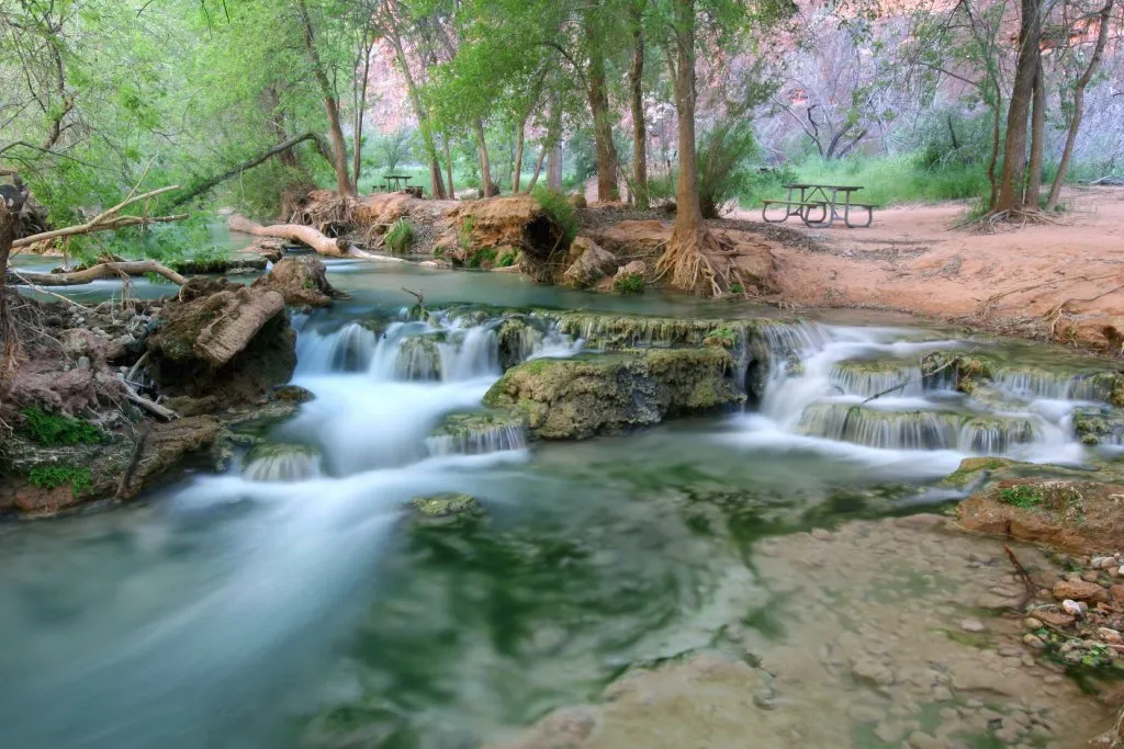 Picnic tables are seen adjacent mini waterfalls that are surrounded by trees and shade.