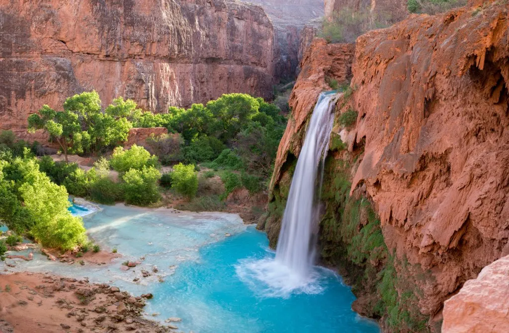 A landscape shot of a waterfall over red rocks pouring into a blue water pond surrounding by more rock and trees.