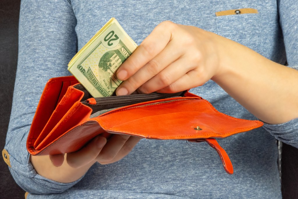 Close-up of a white woman's hands putting twenty dollar bills into an orange wallet. She is wearing a heather gray shirt.
