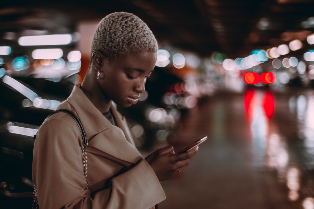 A black woman in a parking garage is looking down at her phone. She has short blonde hair and is wearing a tan trenchcoat.