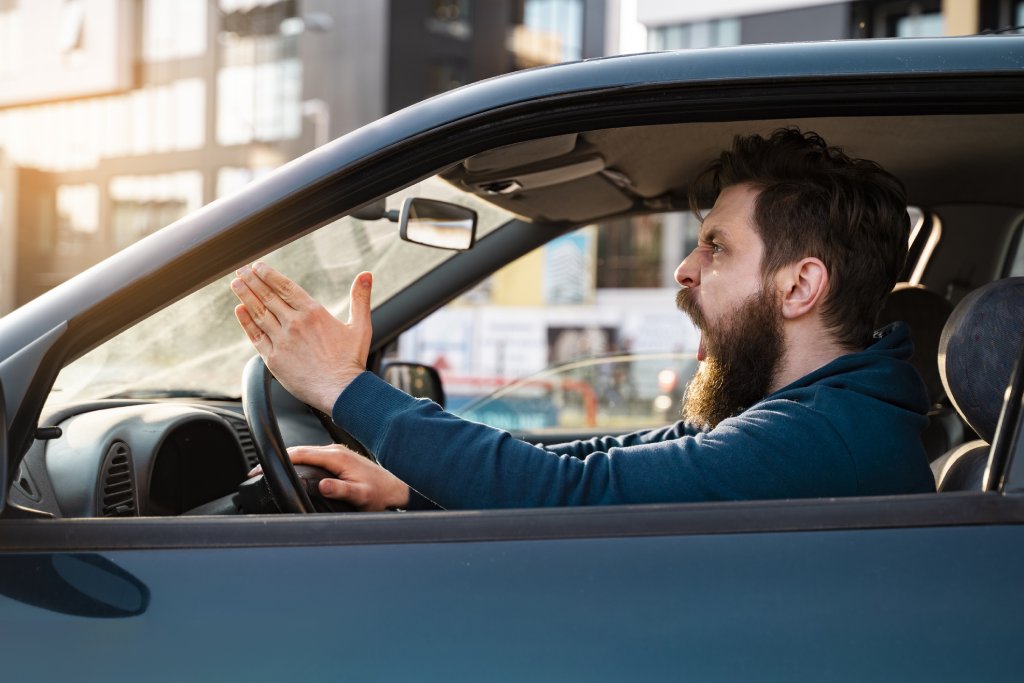 A very angry white guy with a long beard and blue shirt is holding out his hand in disgust. His mouth is open like he's yelling at someone from the driver's seat of his car.