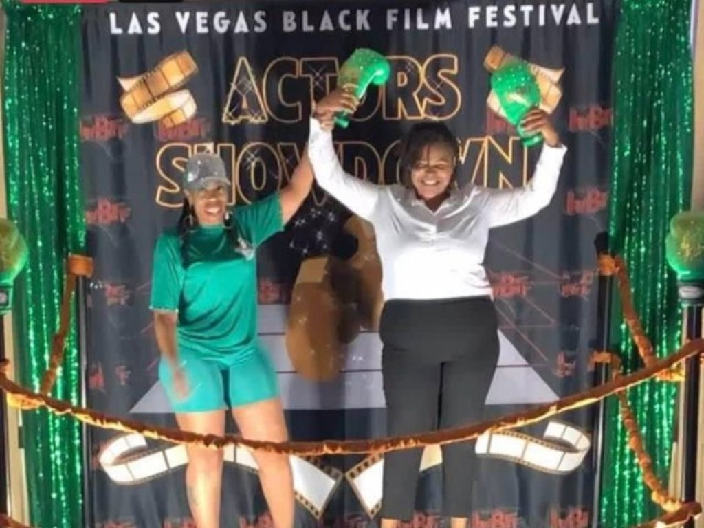 Two women stand in a quasi boxing ring celebrating the win of the woman on the right. She is in black pants and a white top and the one on the left is wearing a green short jumpsuit.